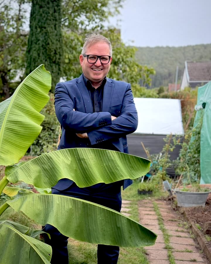 Jörg „Schorni“ Wittmer mit verschränkten Armen im Garten hinter einer Bananenstaude, im Hintergrund die bewaldeten Hügel des Dahner Felsenlands